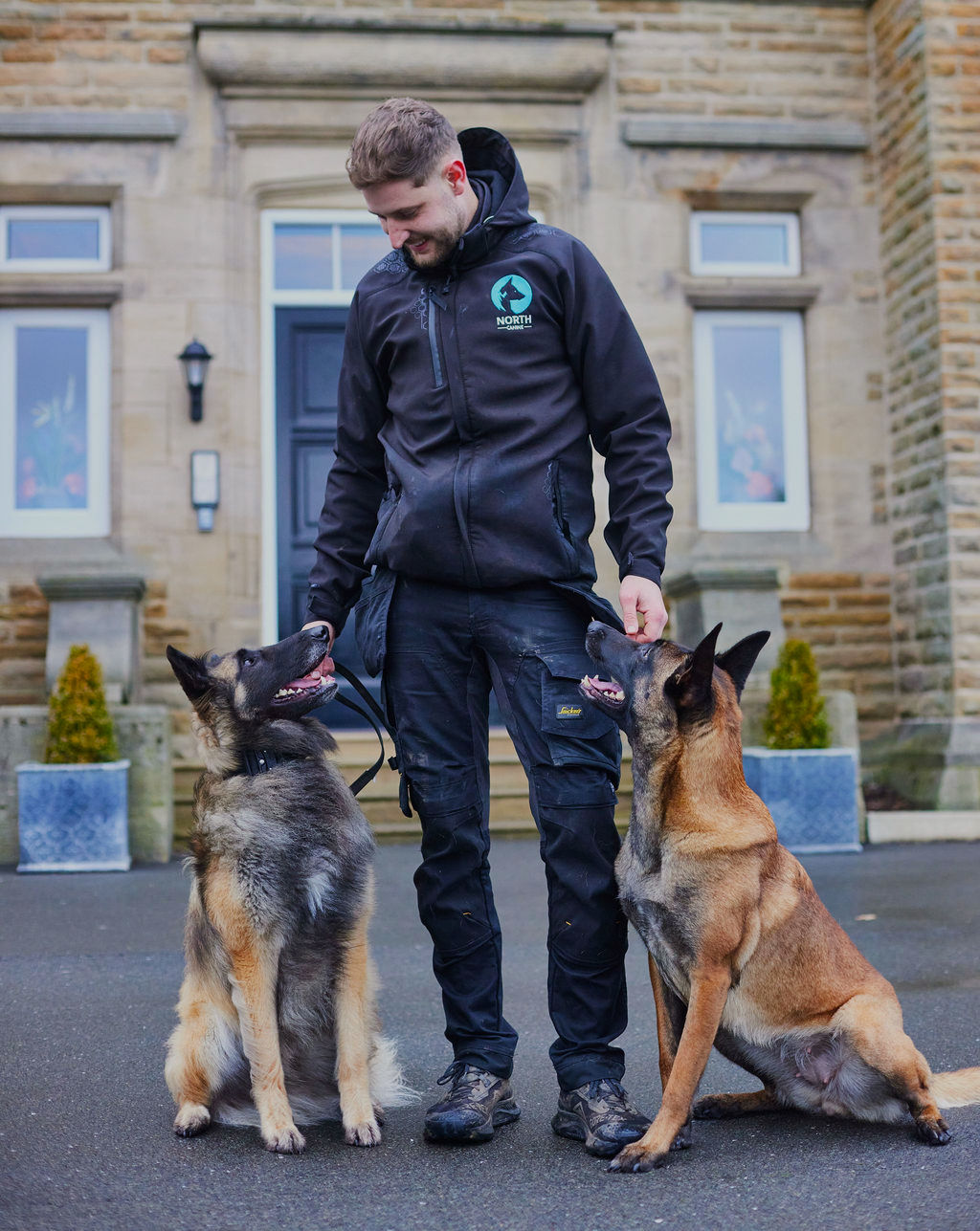 North Canine trainer with two attentive German Shepherd-type dogs outside a stone building